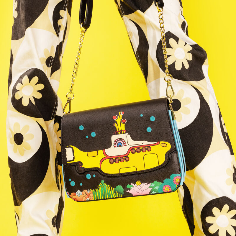 Person standing against a yellow background wearing psychedelic pants and holding the Beatles Yellow Submarine crossbody bag, featuring a yellow submarine against a black background surrounded by bubbles and aquatic plants